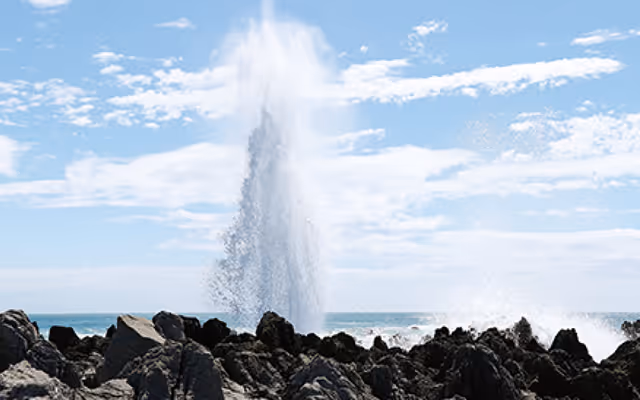 Water spout erupting from rocky coastline under a partly cloudy blue sky.