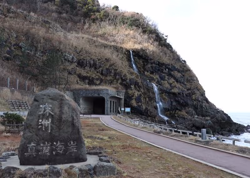 Stone monument with Japanese inscription near a paved path leading to a tunnel at the base of a rocky hill with a waterfall, beside the ocean.