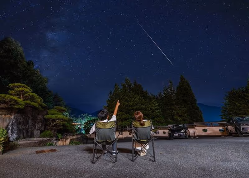 Two people sitting in camping chairs in a parking lot, looking up at a starry night sky with a visible shooting star.