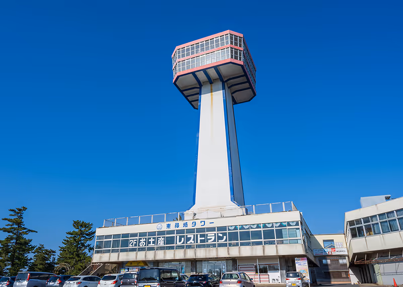 Tall observation tower with a hexagonal glass-walled top section above a building under clear blue sky and parked cars in front.