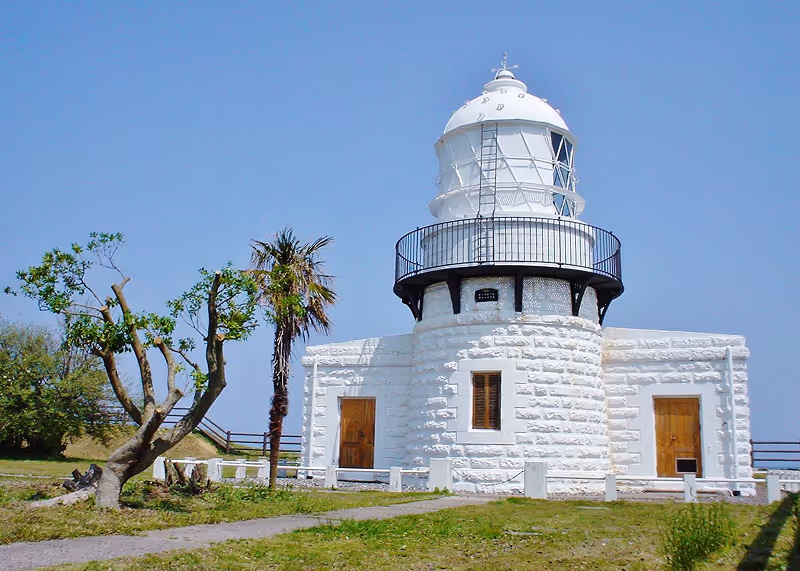 White stone lighthouse with a black railing around the lantern room, set against a clear blue sky with green grass and trees nearby.