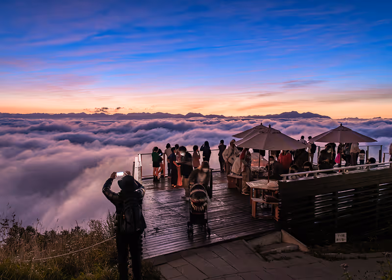 People enjoy a scenic sunset view atop a wooden observation deck overlooking a sea of clouds and distant mountains.