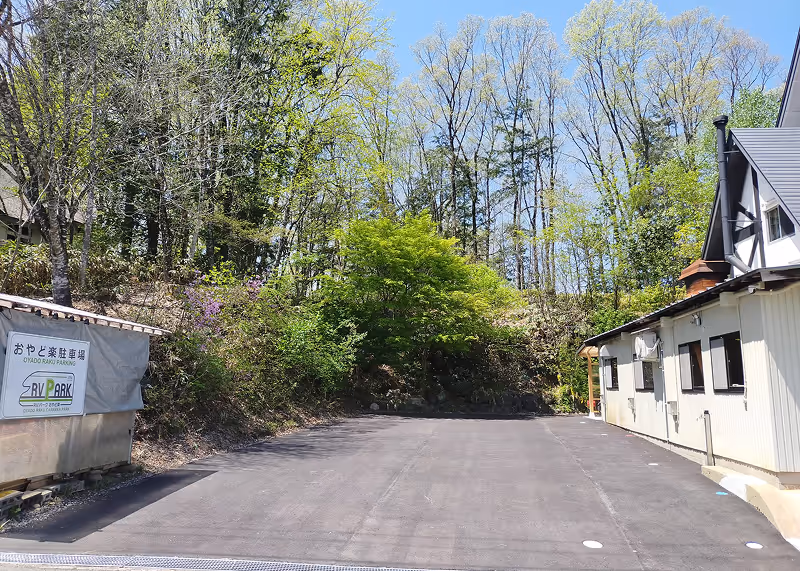 Empty paved parking area bordered by green trees and shrubs on a sunny day next to a white building with a pitched roof.