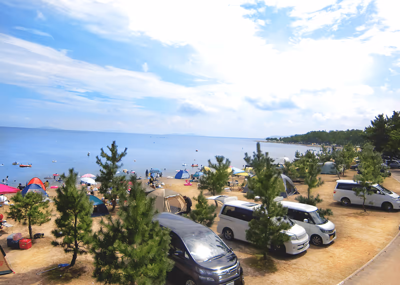Beach campsite with several tents, cars, and small pine trees along a sandy shore beside a calm blue sea under a partly cloudy sky.