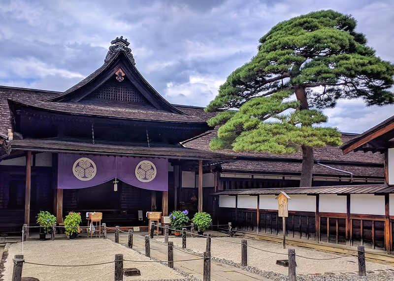Traditional Japanese wooden building with a curved roof, purple hanging banners, and a large manicured pine tree under a cloudy sky.