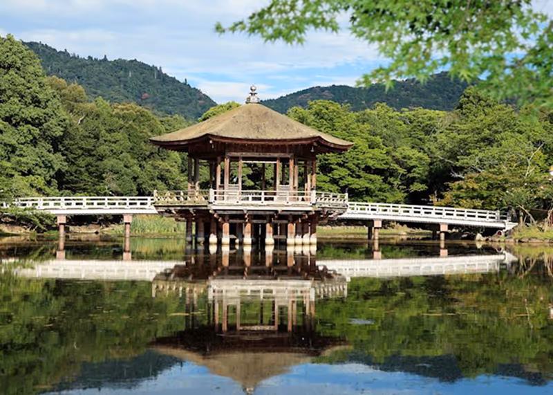 Traditional Japanese gazebo on wooden stilts with white railings reflected in calm pond surrounded by green trees and hills.