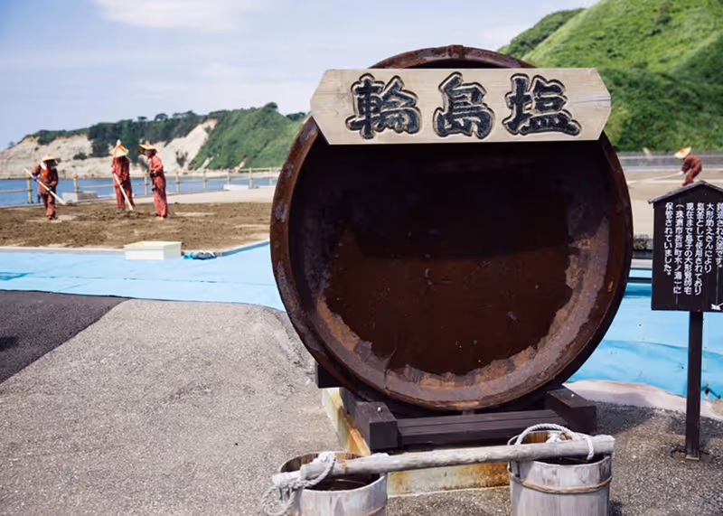 Rusty large barrel with a wooden sign in Japanese characters, workers in traditional attire raking salt on drying beds near a green hillside.