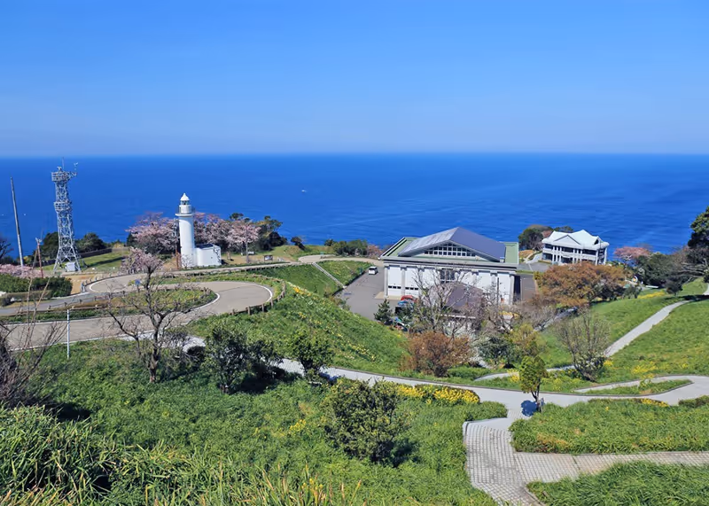 Coastal landscape featuring winding paths, a white lighthouse surrounded by greenery and pink blossoms, buildings near the sea, and a clear blue sky.