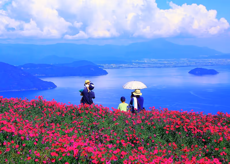 A family of four enjoying a vibrant red flower field overlooking a large blue lake with mountains and a cloudy sky in the background.