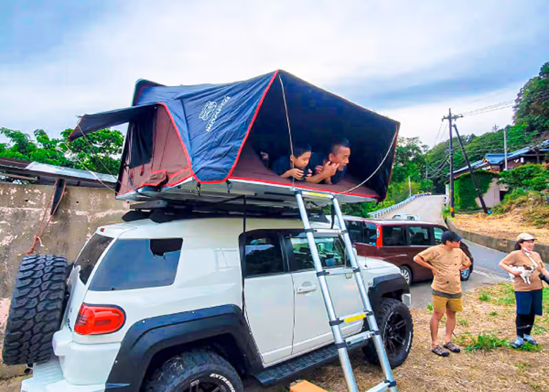 Two people inside a rooftop tent mounted on a white off-road vehicle with a ladder, while two others stand nearby on a roadside campground.