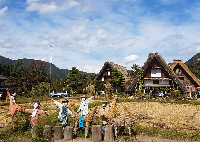 Traditional thatched Japanese houses behind a field with scarecrows and a clear blue sky.