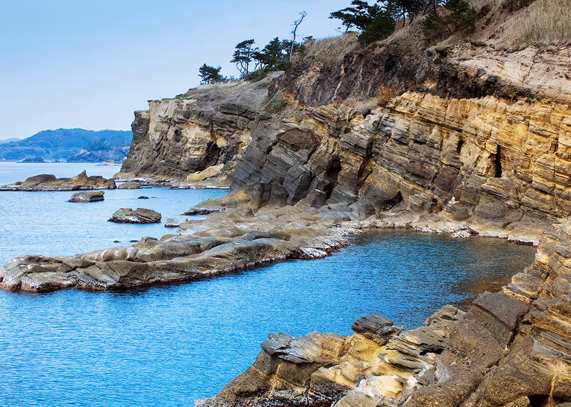 Rocky coastal cliffs with layered sedimentary rock formations beside a calm blue sea under a clear sky.