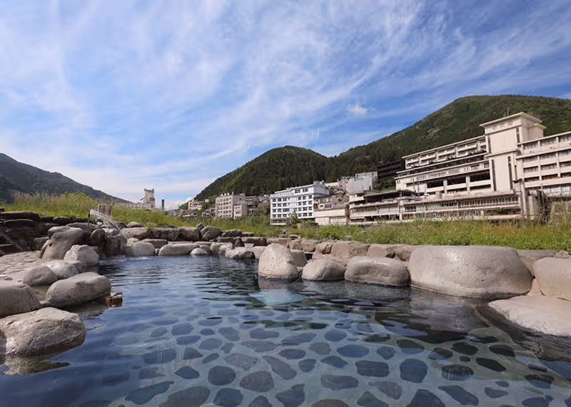 Outdoor stone hot spring bath with clear water, surrounded by large rocks and green hills, with buildings in the background under a blue sky.