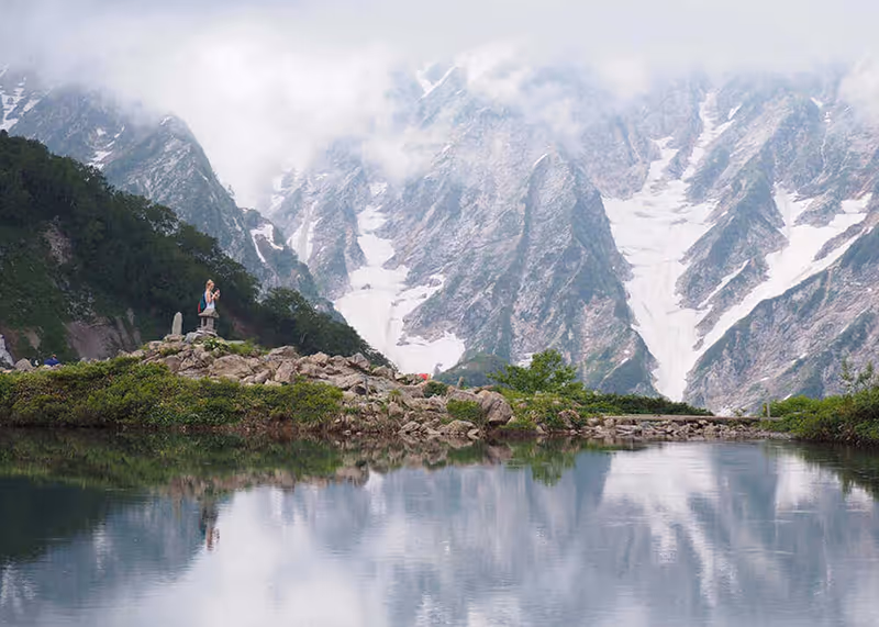 Person taking a photo atop rocky terrain beside a calm pond with snow-capped mountains partially covered by clouds in the background.