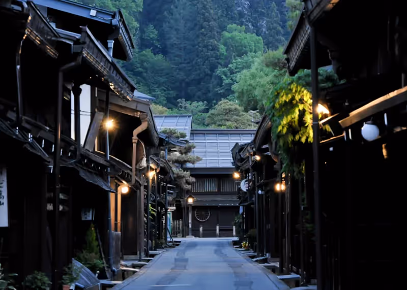 Peaceful narrow street lined with traditional wooden buildings and warm lights at dusk, with forested hills in the background.