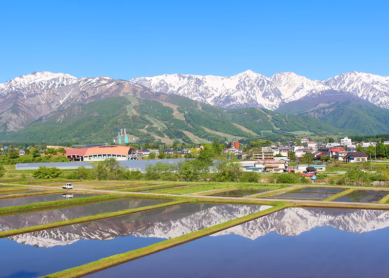 Snow-capped mountains and green hills reflected in flooded rice paddies with a small village in the background under a clear blue sky.