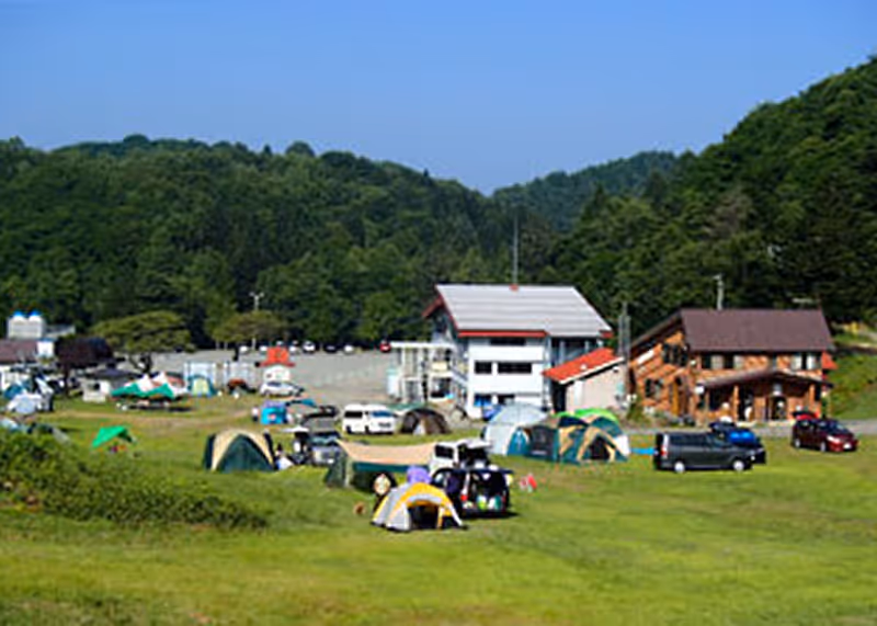 Campsite with multiple tents and parked vehicles on green grass surrounded by forested hills under clear blue sky.