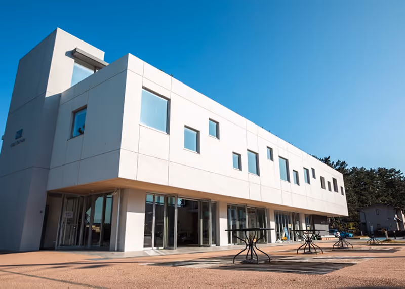 Modern white two-story building with minimalist design, multiple windows, glass doors, and outdoor tables on a sunny day.