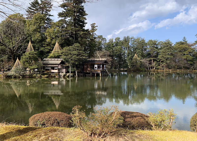Traditional Japanese wooden buildings beside a calm pond surrounded by trees under a partly cloudy sky.