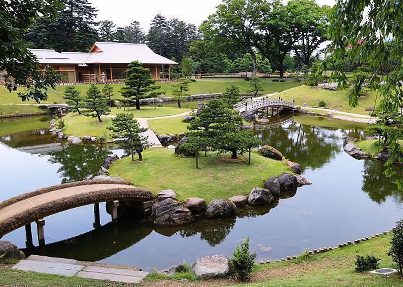 Japanese garden with manicured trees, stone bridges over a pond, and a traditional wooden building in the background.
