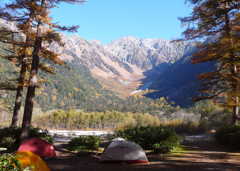 Campsite with tents under trees in front of a mountain range on a clear sunny day.