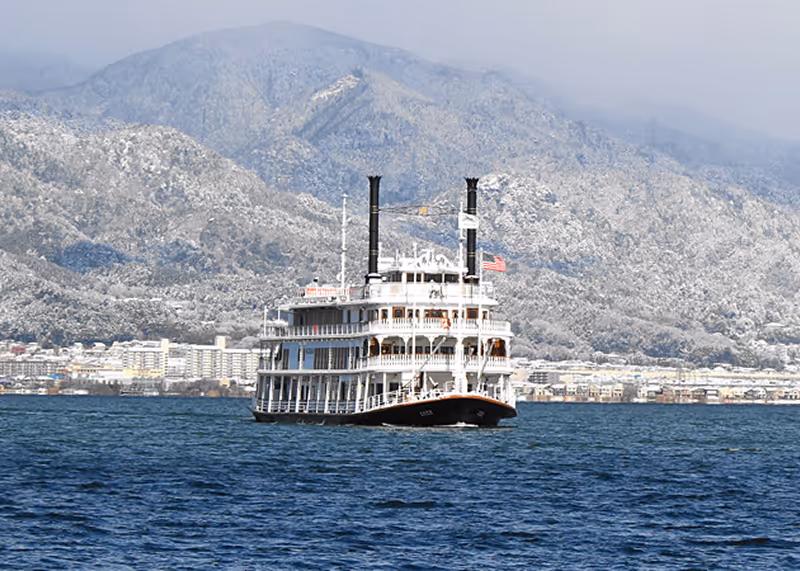 White paddlewheel riverboat cruising on a lake with snow-covered mountains and buildings in the background.