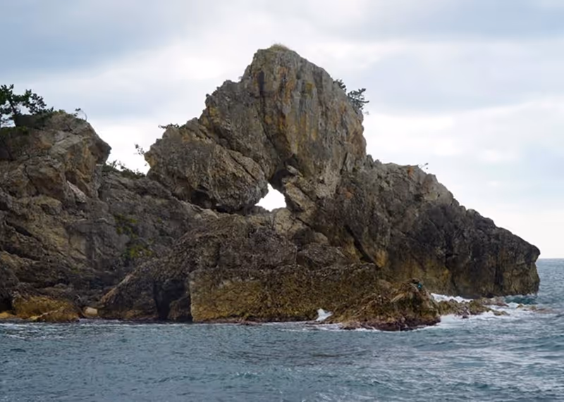 Large rocky formation with a natural arch rising from the ocean under a cloudy sky.