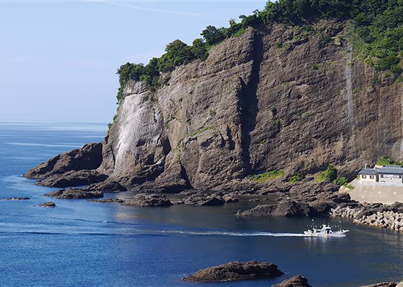Rocky coastal cliff with green foliage above and a small boat moving across the calm blue water below.