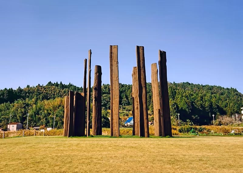 Tall wooden pillars arranged vertically in a grassy park with forested hills and clear blue sky in the background.