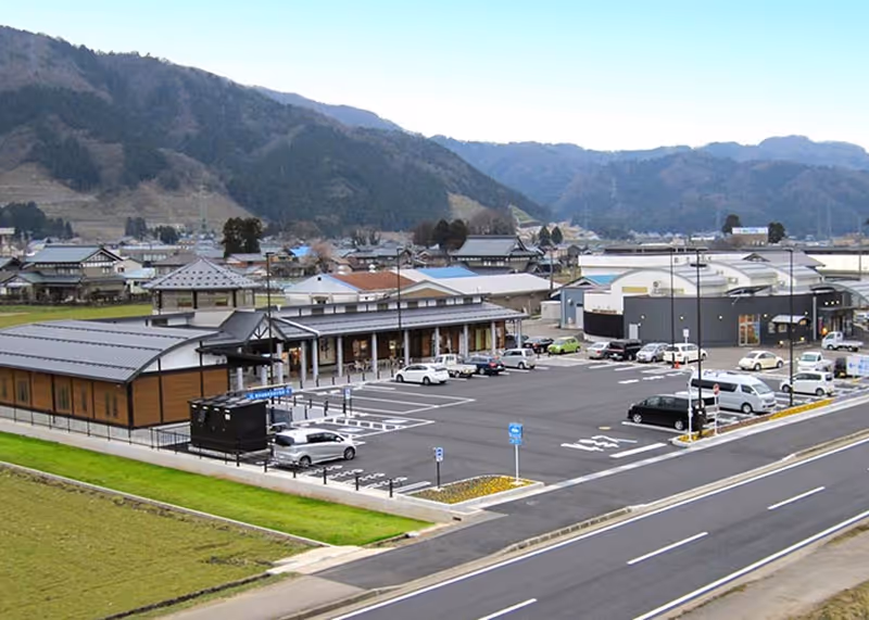 Parking lot with cars adjacent to modern low-rise buildings set against forested mountains and clear sky.
