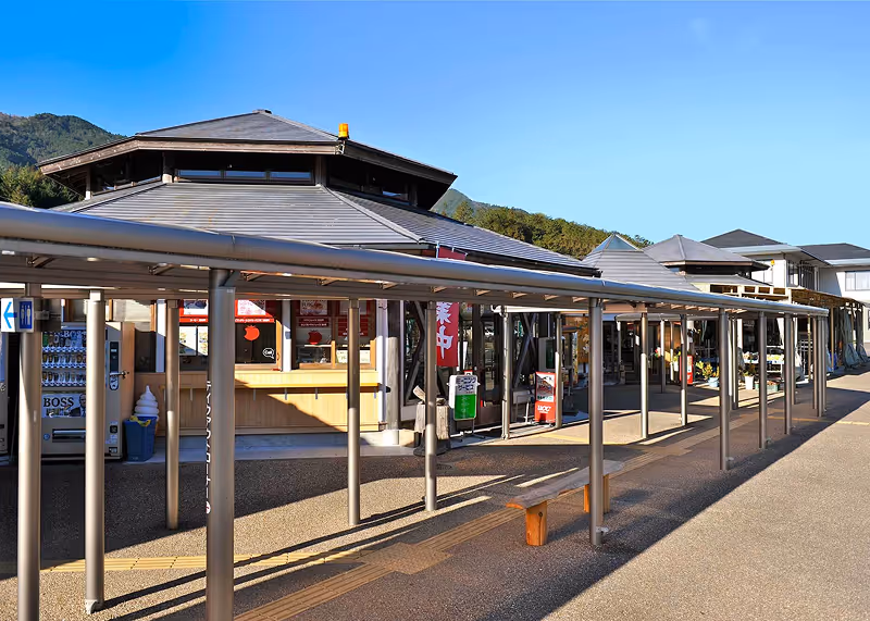 Outdoor walkway with a metal roof supported by pillars leading to a series of buildings under a clear blue sky.
