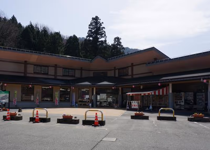 Parking area with safety barriers and orange traffic cones in front of a two-story building surrounded by trees under a partly cloudy sky.