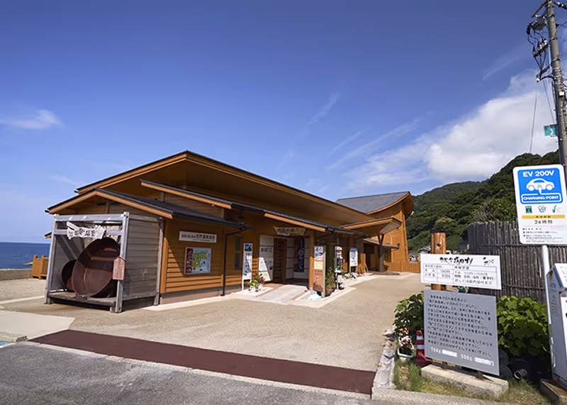 Modern wooden building with a sloped roof near the ocean under a clear blue sky with informational signs and greenery in front.