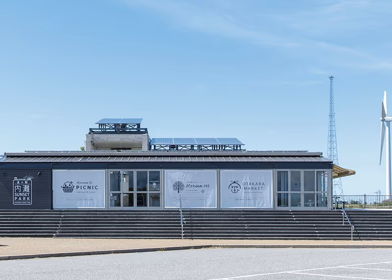 Modern building with solar panels on the roof and banners reading Michinoeki Sunset Park, Picnic, Florica 205, and Otakara Market under a clear blue sky.