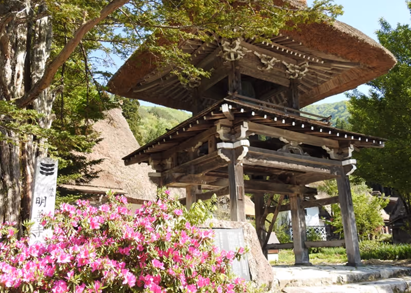 Traditional Japanese wooden temple gate with a thatched roof surrounded by green trees and pink flowers in a sunny garden.