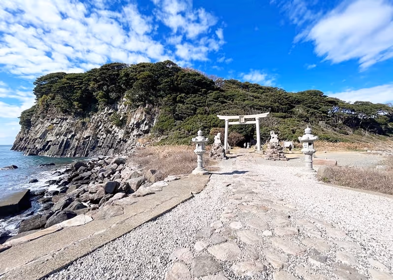 Stone pathway leading to a white torii gate with stone lanterns on either side, set against a coastal cliff covered with trees under a partly cloudy blue sky.