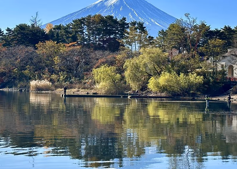 Calm lake reflecting trees and bushes with Mount Fuji snow-capped in the background under clear blue sky.