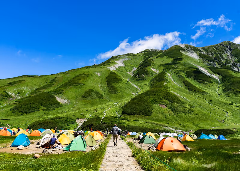 Colorful tents set up on grassy campground at the base of lush green mountains under a clear blue sky.