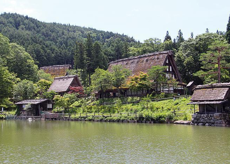 Traditional thatched-roof houses surrounded by lush greenery beside a calm lake with forested hills in the background.