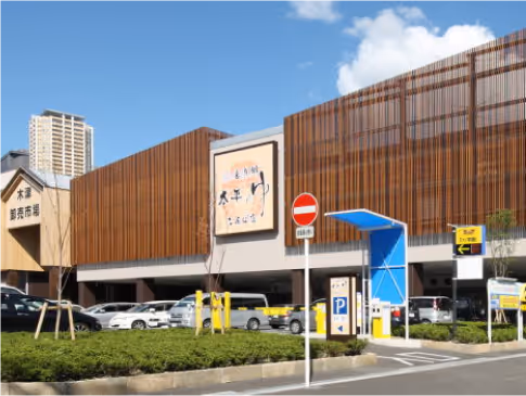 Exterior of Taiheinoyu facility with wooden paneling, parked cars, and a blue parking entrance under a partly cloudy sky.
