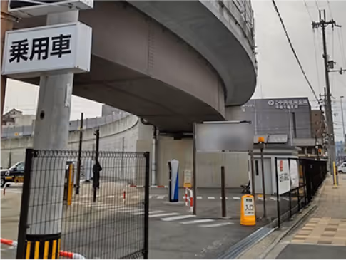 Urban parking lot under a large curved elevated highway with a sign in Japanese and fenced entrance.