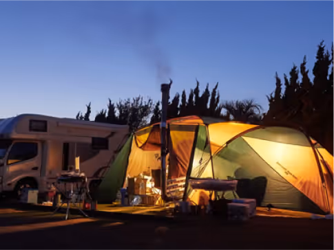 Illuminated camping tent with chimney smoke and a parked RV at dusk in a campsite.