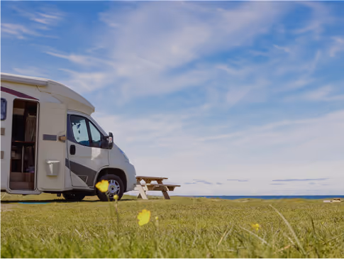 White camper van parked on grassy land near a wooden picnic table under a blue sky with clouds.