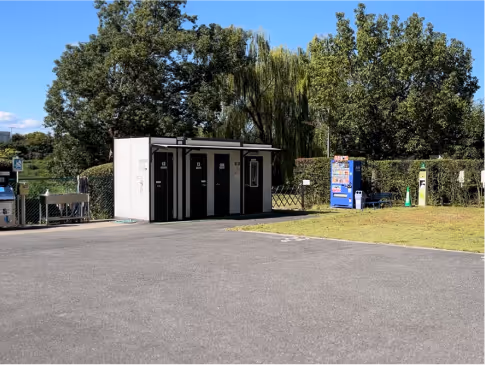 Outdoor restroom facility with three black-and-white doors near a vending machine and surrounded by trees under a clear blue sky.