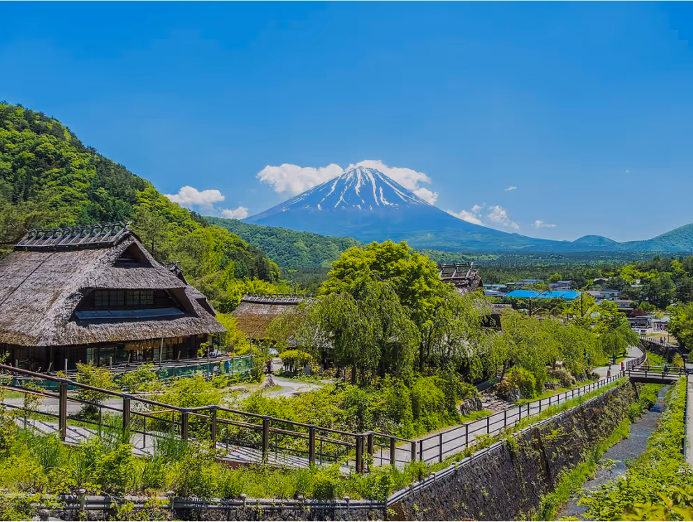 Traditional thatched-roof houses nestled in lush green landscape with Mount Fuji in the background under a clear blue sky.