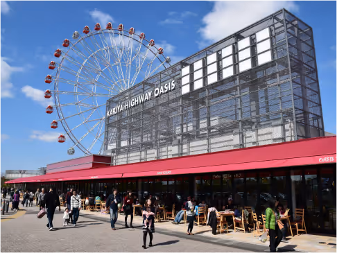Outdoor scene of Kariya Highway Oasis with a Ferris wheel and people walking and sitting at tables under red awnings.