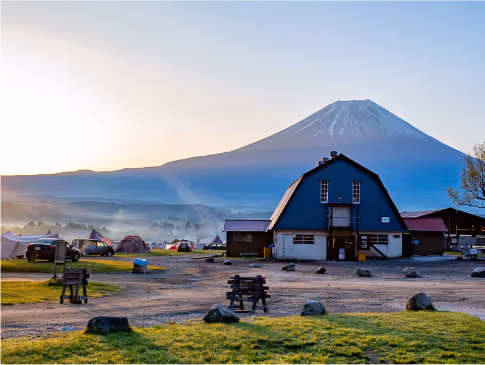 Camping site with tents and a barn-like building in the foreground, with Mount Fuji visible in the background during sunrise.