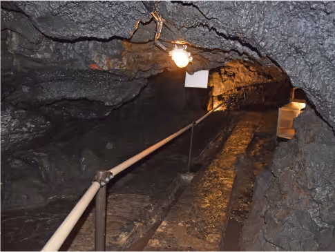 Narrow underground tunnel with rocky walls, a handrail along a stone pathway, and overhead lights.