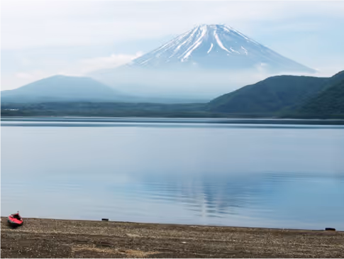 Calm lake with a reflection of snow-capped Mount Fuji under a cloudy sky, with a sandy shore in the foreground.