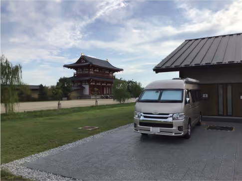 Silver camper van parked next to a modern building with traditional Japanese architecture visible in the background under a cloudy sky.